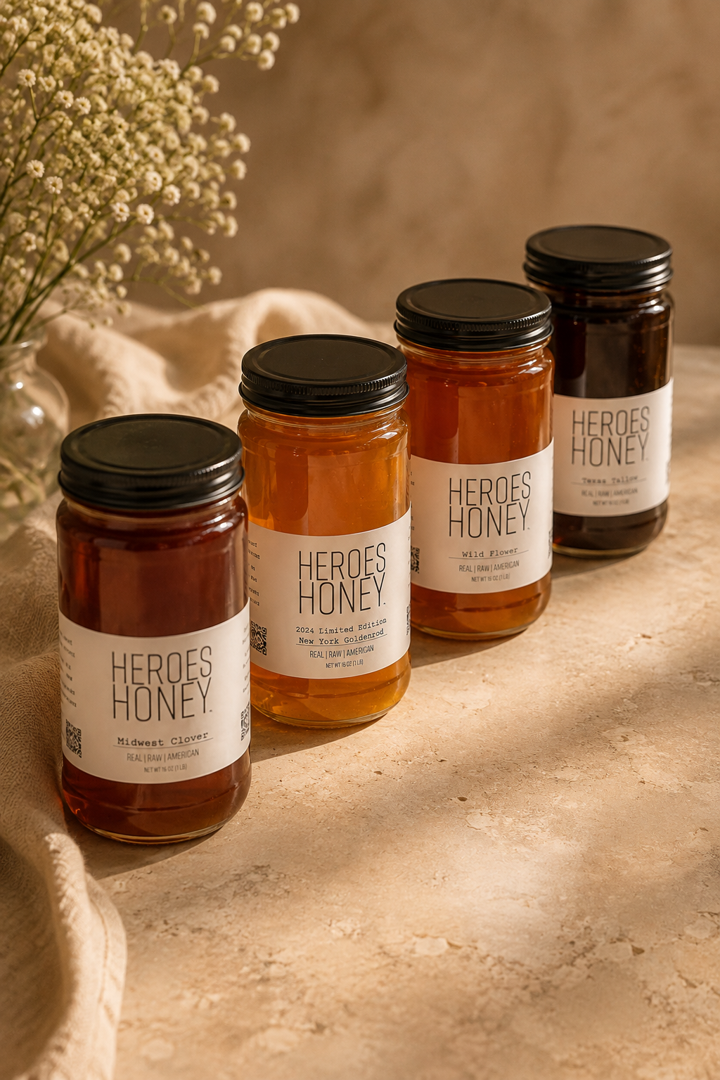 Four Raw Truth Honey jars — Midwest Clover, New York Goldenrod, Wild Flower and Texas Tallow — arranged on a warm stone surface with linen and baby's breath